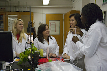 Four female students in a lab stand around a microscope and participate in a photosynthesis activity.