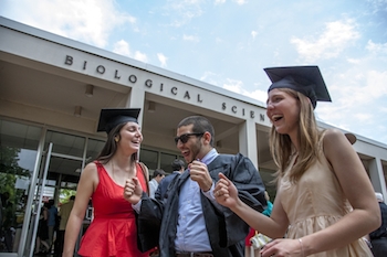 A male in his graduation gown and two females in their graduation camps rejoice outside of the Biosciences building before their reception.