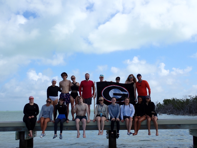 A group of students holding a UGA flag sit on a pier over water.