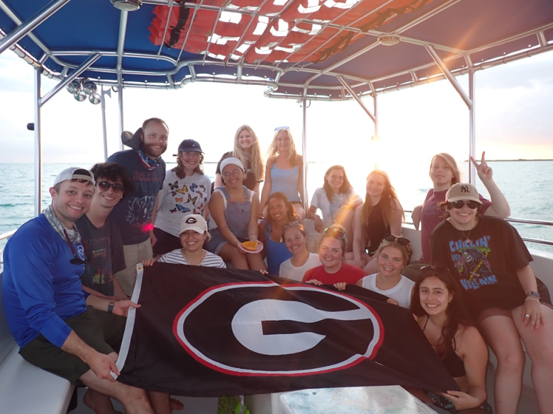 A group of students and instructors on a boat display a UGA flag.
